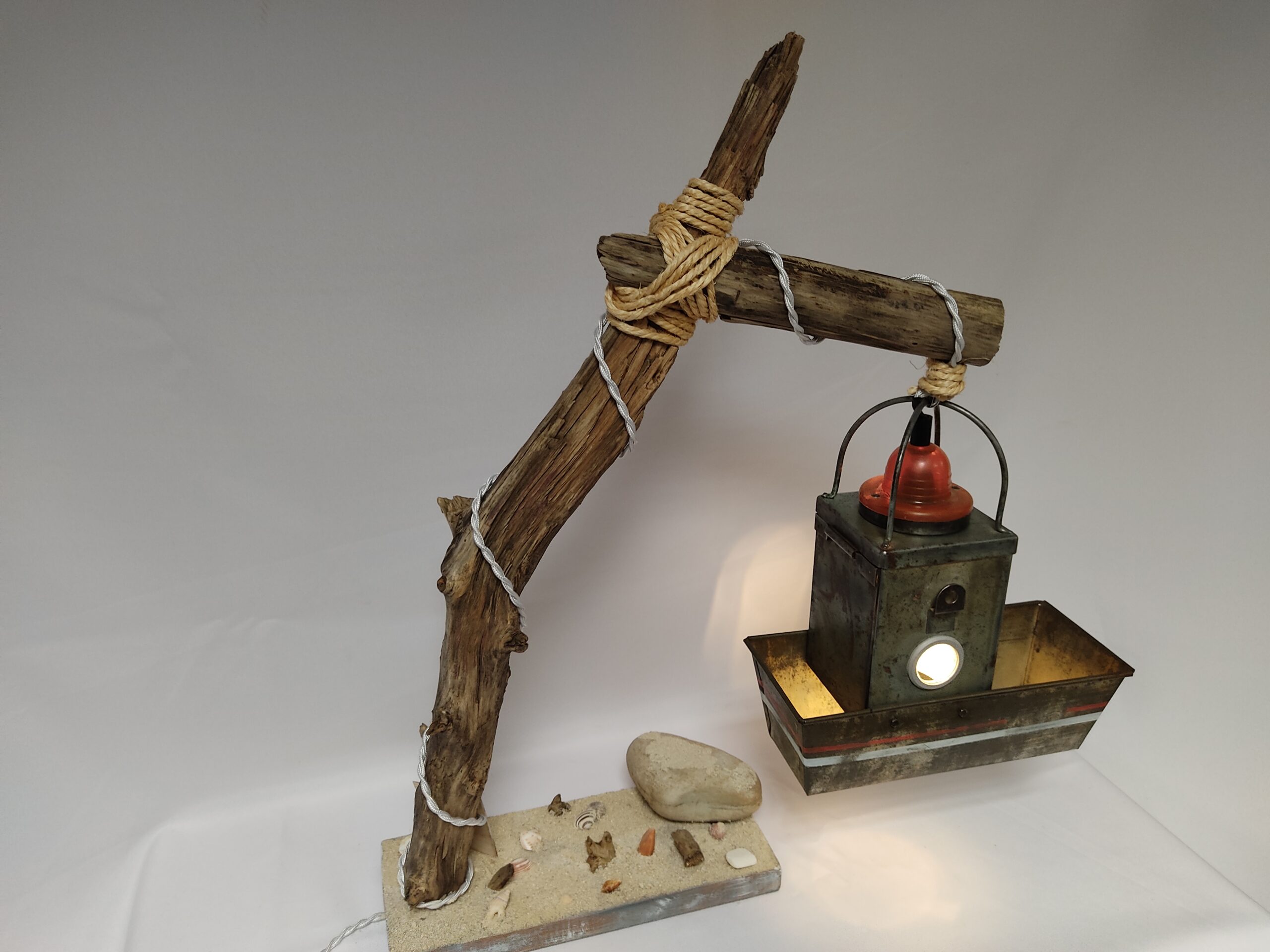 Une lampe bateau de pêche basé sur un moule à cake et une lanterne suspendus à une branche de bois flotté sur un lit de sable et de coquillages